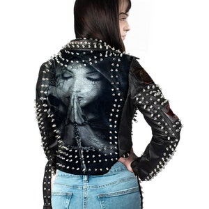 Könnte beinhalten: Schwarze Lederjacke mit silbernen Nieten und einer Grafik einer betenden Frau auf dem Rücken. Die Jacke hat einen klassischen Biker-Stil mit einem Reverskragen, einem asymmetrischen Reißverschluss und Nieten an Schultern und Ärmeln.