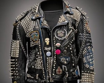 Könnte beinhalten: Schwarze Lederjacke, stark verziert mit silbernen Nieten und Stacheln. Die Jacke hat verschiedene Aufnäher, Anstecknadeln und Ketten, mit einem Reißverschluss und einem Gürtel. Die Ärmel und Schultern sind mit Metallnieten und Stacheln bedeckt. Auf dem Ärmel steht das Wort "MISANTHROPY".