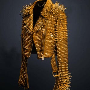 Puede incluir: Chaqueta de cuero dorada cubierta de pequeños tachuelas redondos y púas más grandes y puntiagudas. La chaqueta tiene un estilo motero clásico con cierre de cremallera, solapas con muescas y cinturón en la cintura. Una prenda llamativa.