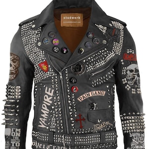 Op de afbeelding: Zwarte leren jas rijkelijk versierd met zilveren studs en patches. Met de tekst "VAMPIRE", "PAIN GANG" en "DEATH IN METAL". Bevat een schedelafbeelding en diverse decoratieve spelden. Een opvallend kledingstuk.