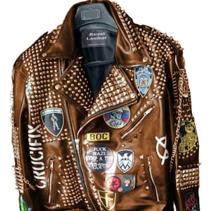 May include: Brown leather biker jacket adorned with metal studs and patches. The jacket has a zip closure and multiple pockets. Patches include the text "CRUCIFIX" and "FUCK NAZI SYMP A TH". The jacket is hanging on a hanger.