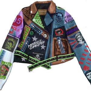 Könnte beinhalten: Eine schwarze Lederjacke mit einer Vielzahl von bunten Patches und Nieten. Die Patches zeigen Bandlogos, politische Figuren und Slogans. Die Jacke hat einen grünen Reißverschluss und einen braunen Kragen.
