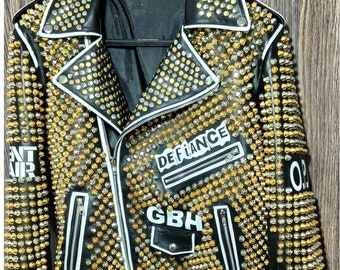 Pode incluir: Jaqueta de couro preta ricamente adornada com tachas douradas e prateadas. Apresenta acabamentos brancos, zíperes e a palavra "DEFiance" em um patch. A jaqueta tem um estilo motociclista clássico com cinto.