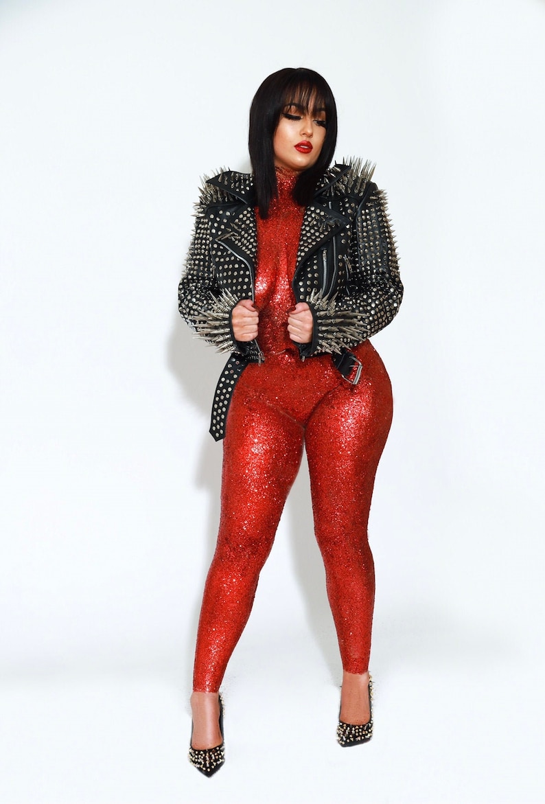 May include: A woman in a red sequined jumpsuit and a black leather jacket with silver spikes. The jacket is belted at the waist. She is wearing black heels with silver studs. The background is white.