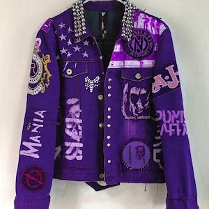 以下が含まれることがあります： 襟にシルバースタッズが付いた紫色のデニムジャケット。ジャケットには、星、テキスト、イラストなど、さまざまなパッチとグラフィックが施されています。袖にはテキストとパッチがあります。ジャケットはボタンで留められ、白いハンガーに掛けられています。