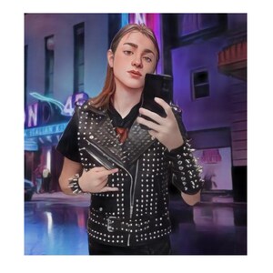 Könnte beinhalten: Eine schwarze Lederweste mit silbernen Nieten und Stacheln. Die Weste wird über einem schwarzen T-Shirt getragen. Ein silbernes Armband befindet sich am Handgelenk. Der Hintergrund zeigt ein Neon-Schild und eine Stadtstraße.