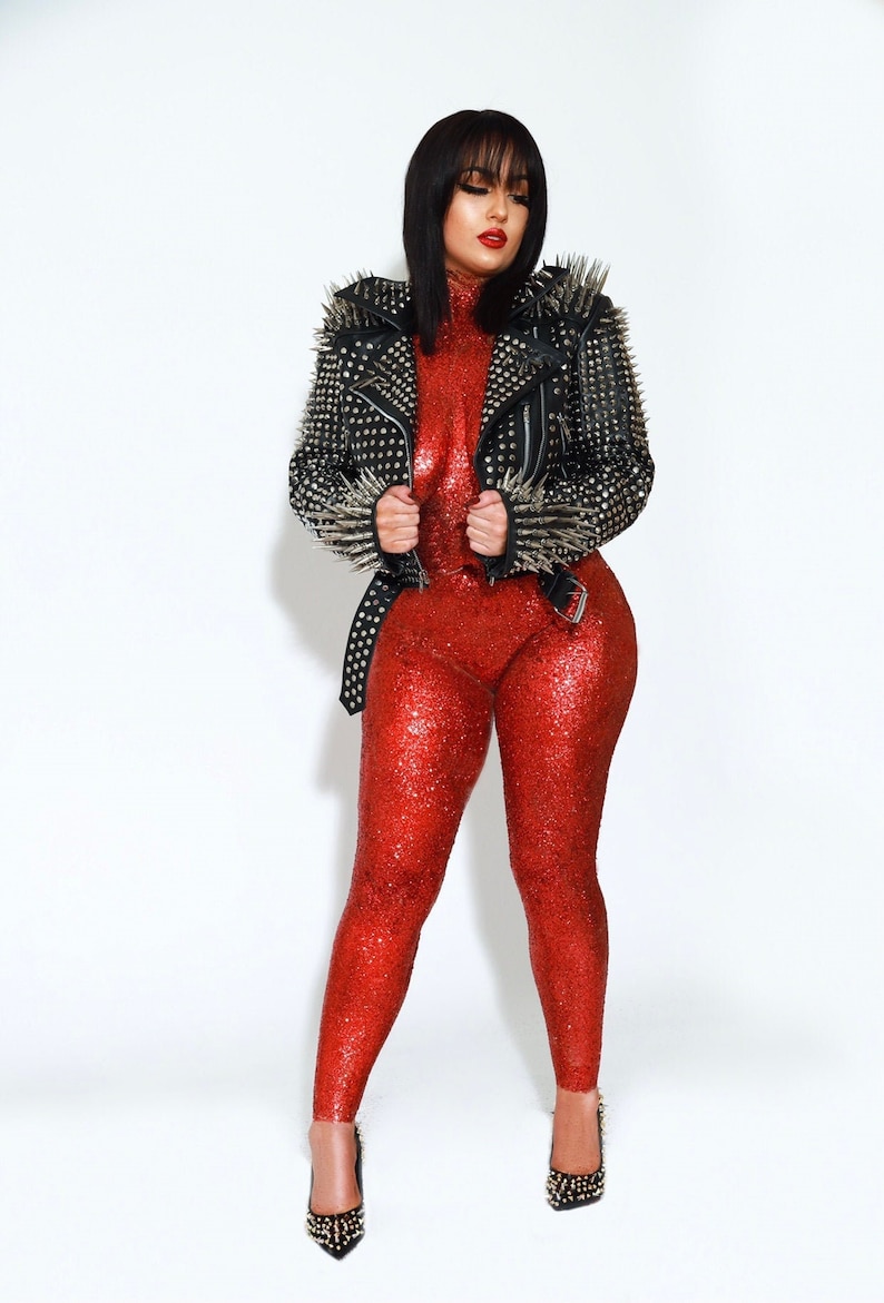 May include: A woman in a red sequined jumpsuit and black studded jacket. The jacket has silver spikes and a belt. She is wearing black heels with gold studs. The background is white.