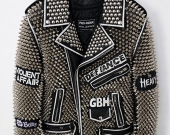 Può includere: Una giacca da moto in pelle nera riccamente decorata con borchie e punte in metallo argentato. La giacca presenta revers con bordini bianchi, tasche con cerniera, una cintura con fibbia e toppe con il testo Violent Affair, The Bomb, Defiance e GBH.