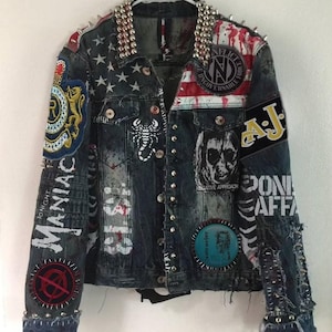 Peut inclure: Une veste en jean bleue avec divers patchs, dont un drapeau américain, un scorpion et le texte "MANIAC" et "NEGATIVE APPROACH". La veste est recouverte de clous argentés et a un aspect vieilli.