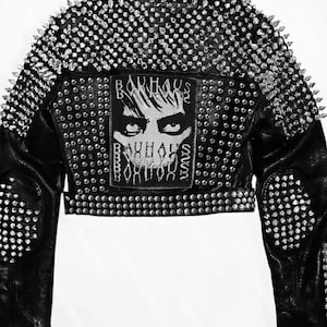 May include: Black leather jacket heavily embellished with silver studs and spikes. The back features a Bauhaus patch with a stylized face and the band's name. The jacket has a cropped design and a classic biker style.