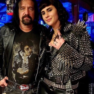 May include: Two people are shown in a photo. One person wears a black t-shirt with an image of Elvis Presley and a belt buckle that says "Elvis". The other person wears a black leather jacket covered in silver spikes.