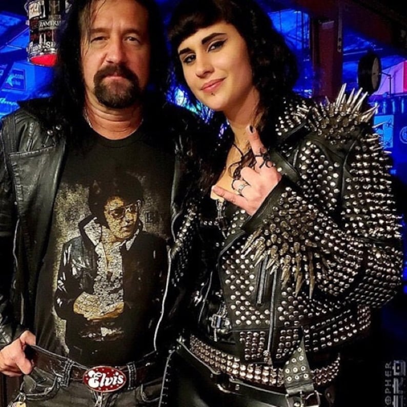 May include: Two people are shown in a photo. One person wears a black t-shirt with an image of Elvis Presley and a belt buckle that says "Elvis". The other person wears a black leather jacket covered in silver spikes.
