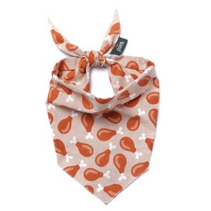 May include: Tan dog bandana with a repeating pattern of cartoon drumsticks in shades of brown and white. The bandana is tied at the top, with a small green tag that reads "paws".