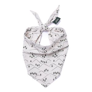 May include: Cream-colored bandana with a mountain and tree pattern. The design features gray mountain ranges and small trees. The bandana is tied at the top, with a small green tag attached. This accessory is suitable for pets.