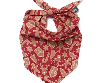 May include: Red dog bandana with a repeating pattern of gingerbread men, houses, trees, and candy canes in tan. The bandana is tied at the top, with a small green tag attached.