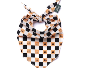 May include: A triangular pet bandana featuring a black and orange checkered pattern. Each orange square contains a small black jack-o'-lantern face. The fabric is tied at the top and includes a small green rectangular tag with the text The Paws.
