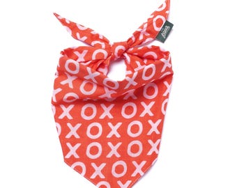 May include: An orange dog bandana with a white "XOXO" pattern. The bandana is tied at the top, with the ends forming a bow. A small green tag is attached to the top right corner.
