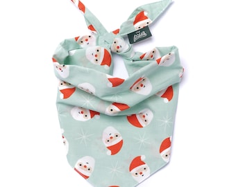 May include: A mint green dog bandana with a repeating pattern of Santa Claus faces and white starbursts. The bandana is tied at the top and features a small tag with the text "The Paws".