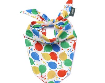 May include: A white dog bandana with a colorful balloon pattern in red, blue, green, and yellow. The bandana is tied at the top and features a small tag with text. The design includes balloon shapes and curly ribbon.