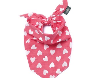 Op de afbeelding: Een roze bandana met een wit hartpatroon, geknoopt. De bandana heeft een label met de tekst "the paws". Dit accessoire voor huisdieren is ontworpen om om de nek te worden gedragen.