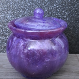 May include: A round, purple resin jar with a matching lid featuring a swirling, pearlescent finish. The container has a wide, curved body and a small, spherical knob on top of the lid, showcasing a glossy, marbled texture throughout the entire piece.