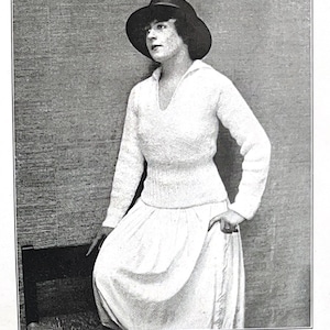 May include: A white knitted sweater with a sailor collar and long sleeves. The sweater is fitted at the waist and has a loose, flowing skirt. The model is wearing a black hat.