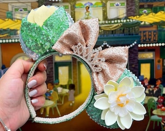 May include: A headband featuring two ears covered in green sequins, a large rose gold sequined bow in the centre, a silver-toned tiara accent on the bow, and a large white water lily flower attached to the right ear with yellow centre details.