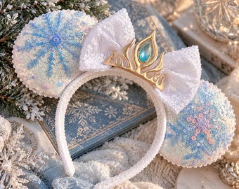 May include: A white headband featuring two circular ears covered in iridescent sequins and blue snowflake embroidery. A central white sequined bow is adorned with a gold-toned metal crown piece containing a single blue teardrop-shaped gem.