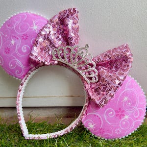 May include: Pink and white mouse ears with a sparkly pink bow and a silver tiara. The ears have a white swirl pattern.