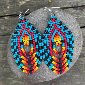 May include: A pair of beaded teardrop earrings. The earrings feature a vibrant pattern of black, red, blue, orange, and yellow beads. The earrings are made with a beaded design and have silver-colored ear wires.