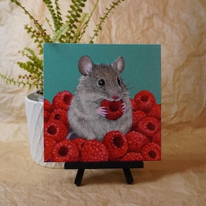 May include: A square painting on canvas featuring a small gray mouse with pink ears and paws, holding a bright red raspberry. The mouse is surrounded by a pile of red raspberries against a solid teal background. The painting sits on a black easel.