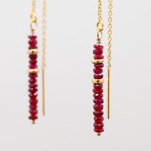 May include: A pair of gold dangle earrings with a long chain and a red ruby bead drop. The earrings are displayed on a gold stand.