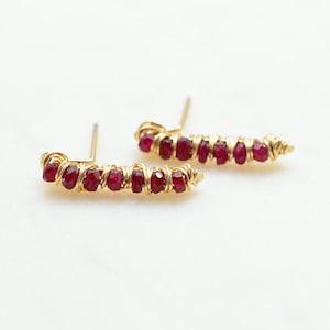 May include: A pair of gold earrings with a curved shape, each earring is adorned with a row of small, red ruby stones wrapped in gold wire.