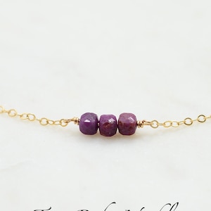 May include: A delicate gold chain necklace with three small, faceted ruby beads. The necklace is on a white background and the text "Tiny Ruby Necklace" is written below the necklace.