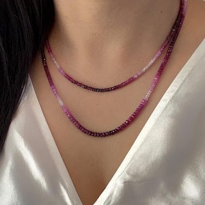 Peut inclure: Deux colliers superposés avec de petites perles rondes roses et violettes multicolores. Les colliers sont portés sur un haut en satin blanc. Les perles passent du rose clair au violet foncé.