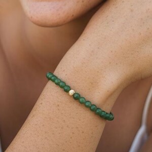 May include: A green beaded bracelet with a gold accent bead is worn on a wrist. The person wearing the bracelet is smiling and looking off-camera. The background is blurred, with trees and natural light.