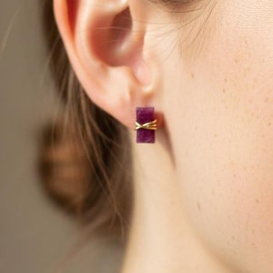May include: Close-up of a person wearing rectangular, dark purple earrings. The earrings are accented with gold-coloured wire. The person is smiling, with their hair pulled back. The earrings are a jewellery accessory.