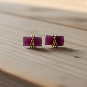 May include: A pair of rectangular earrings with a deep magenta hue. Each earring features a magenta stone wrapped with delicate gold-colored wire. The earrings are displayed on a light-colored wooden surface.