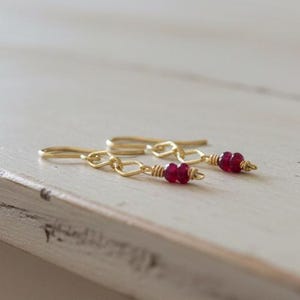 May include: A pair of gold-colored earrings with a chain design and small, red gemstone beads. The earrings are displayed on a white, distressed wooden surface. The jewelry is a delicate accessory.
