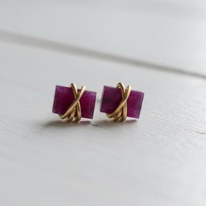 May include: A pair of square stud earrings with a deep magenta hue. Each earring features a magenta square wrapped with gold-colored wire. The earrings are set against a white wooden surface.
