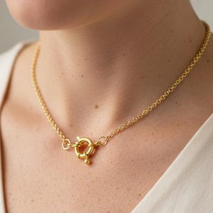 May include: A gold-colored necklace with a circular chain and a decorative clasp. The necklace is worn on a person wearing a cream-colored top. The clasp is the focal point of the necklace.