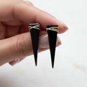 May include: A pair of black, triangular earrings with gold wire accents. The earrings are held in a hand against a white background. The earrings have a geometric design and a modern aesthetic.