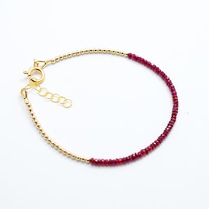 May include: Delicate gold beaded bracelet with a section of small, deep red gemstones. The bracelet has a gold clasp and an adjustable chain. The jewelry is set against a white background.