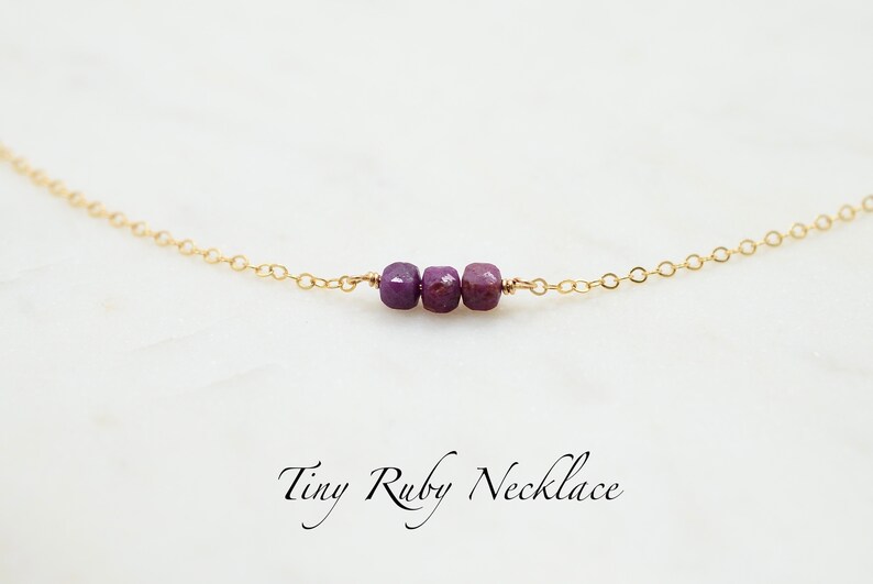 May include: A delicate gold chain necklace with three small, faceted ruby beads. The necklace is on a white background and the text "Tiny Ruby Necklace" is written below the necklace.
