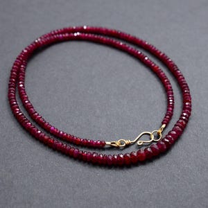 May include: A necklace composed of small, faceted, deep red gemstones. The necklace is secured with a gold-colored clasp. The gemstones are strung together, forming a delicate, layered design. The necklace is displayed against a dark gray background.
