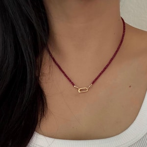 May include: A delicate necklace featuring a strand of small, dark red beads. The necklace has a gold-colored clasp in the center. The necklace is worn around the neck.