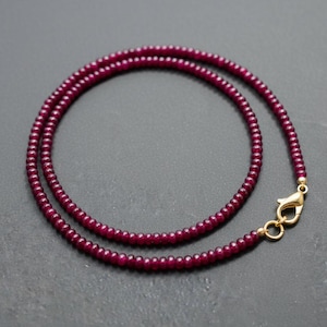 May include: A delicate necklace featuring small, round, deep red beads. The beads are strung together to form a necklace with a gold-colored clasp. The necklace is displayed on a dark gray surface.