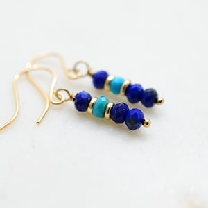 May include: A pair of gold-colored dangle earrings. Each earring features a series of blue and turquoise beads, interspersed with gold-colored accents. The earrings are designed with a simple, elegant style, perfect for everyday wear.
