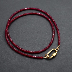 May include: A delicate necklace composed of small, deep red beads. The necklace is secured with a gold-colored clasp. The beads are evenly sized and strung together to form a continuous loop. The necklace is displayed on a dark surface.