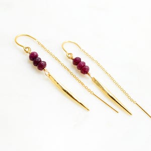 May include: Gold-tone threader earrings with three deep red beads and a gold bar pendant. The earrings have a delicate chain and are displayed on a white surface. The jewelry is a simple, elegant design.